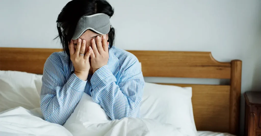 Image of a tired woman sitting in bed covering her face while wearing a sleep mask